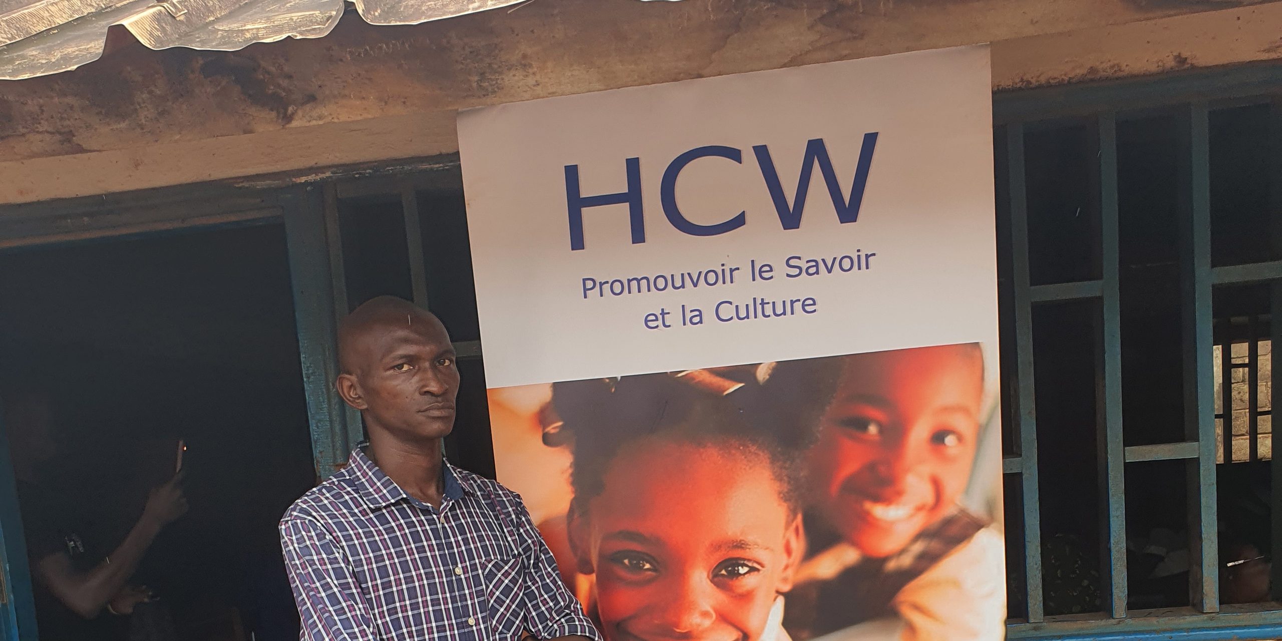 Un kakémono HCW déployé devant un bâtiment scolaire ; on y lit « HCW — Promouvoir le Savoir et la Culture ». Un homme se tient à côté.